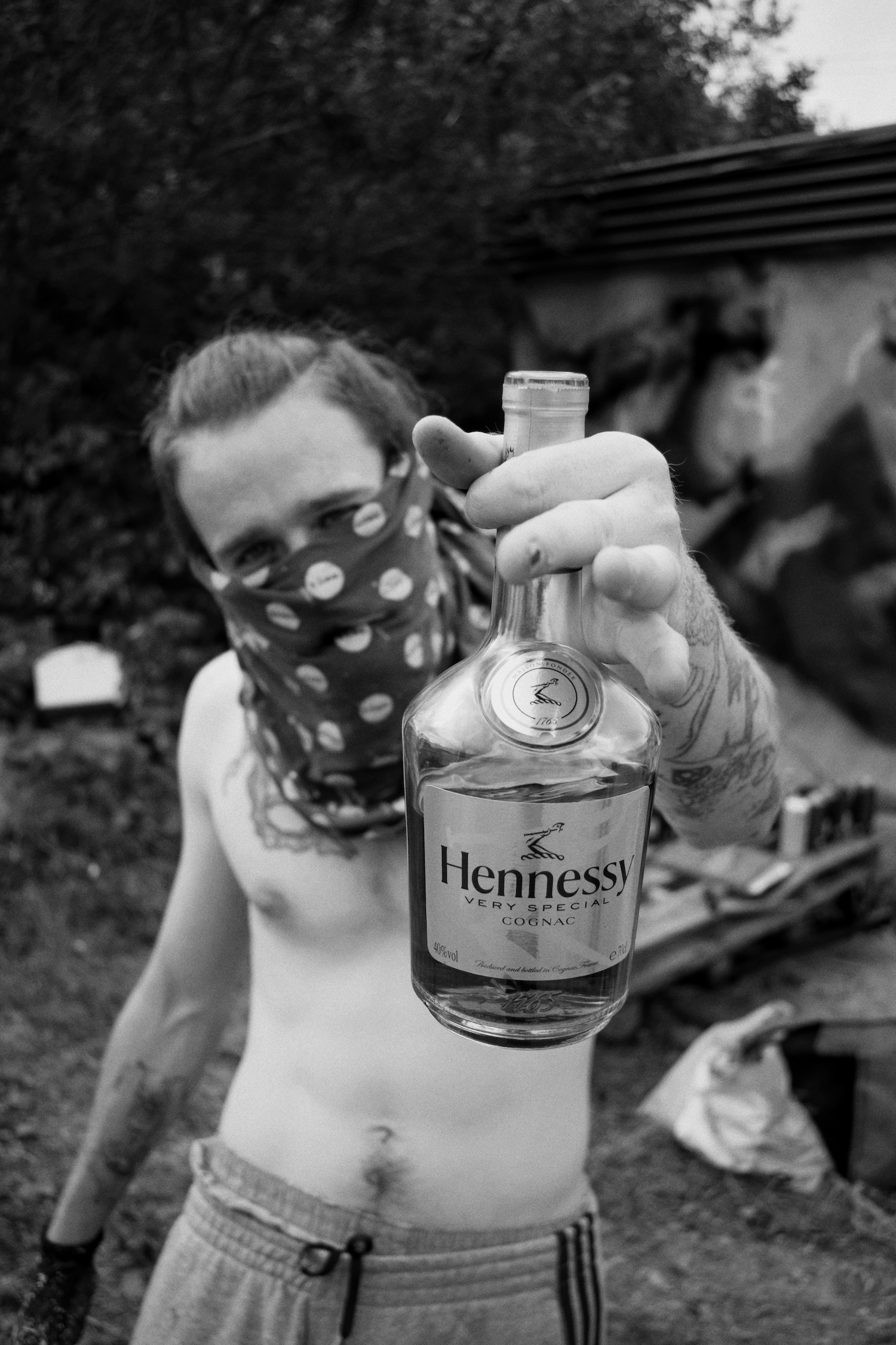 a man with his face covered holds a bottle of cognac to the camera